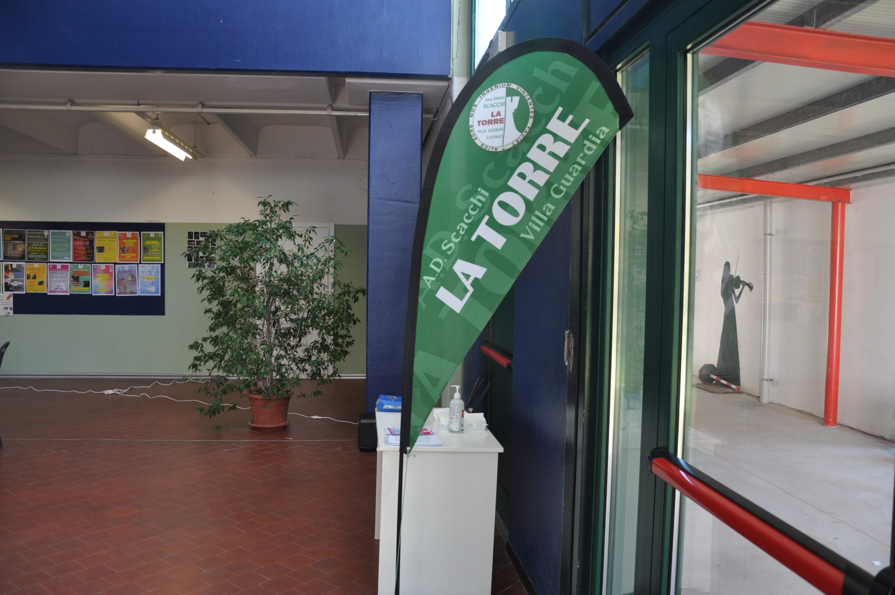 Vertical banner with 'LA TORRE' text in a green design, placed near an entrance with a potted plant and interior walls decorated with posters.