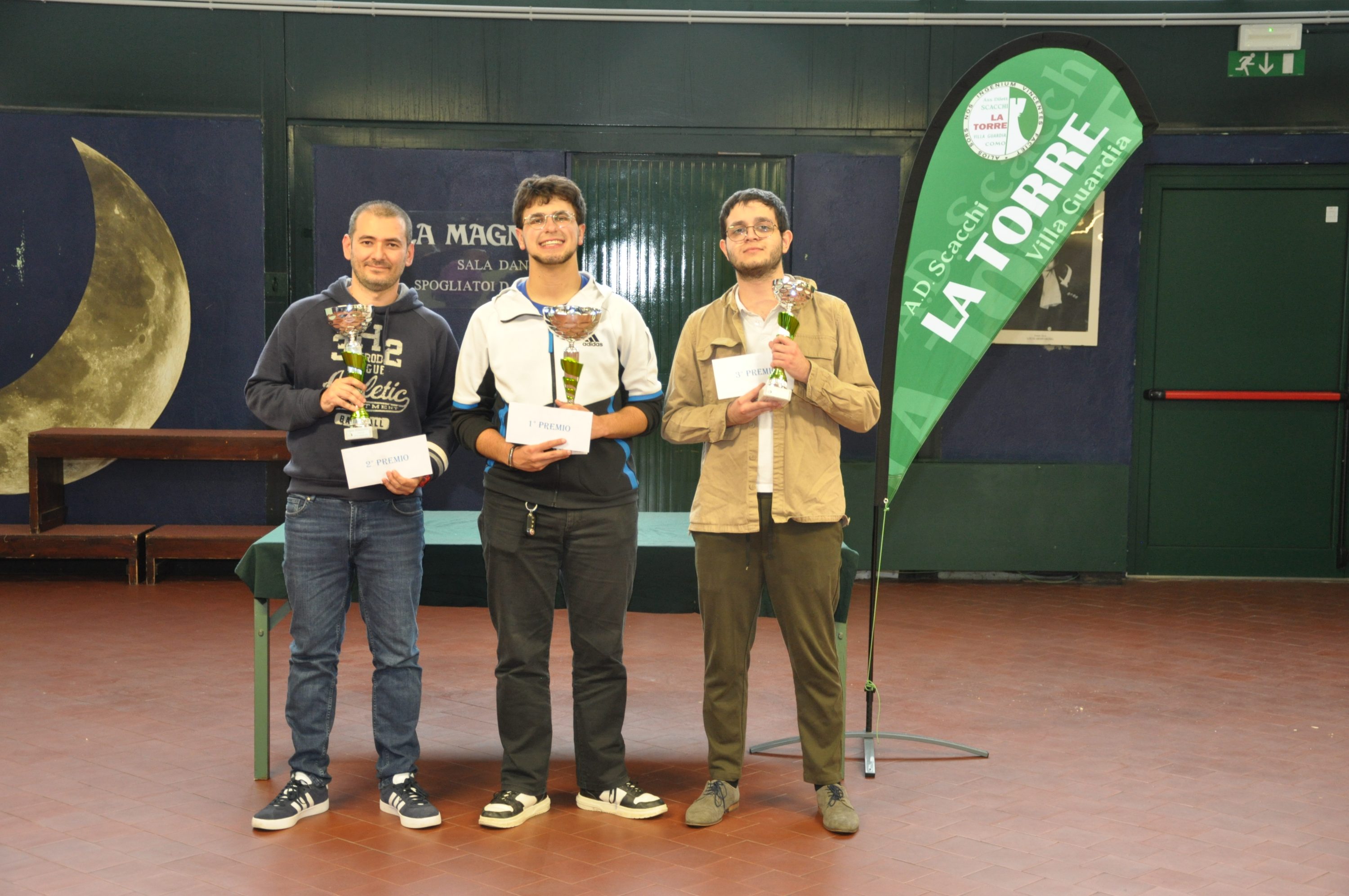 Three individuals standing on a stage holding trophies and certificates, with a banner reading 'LA TORRE' in the background.