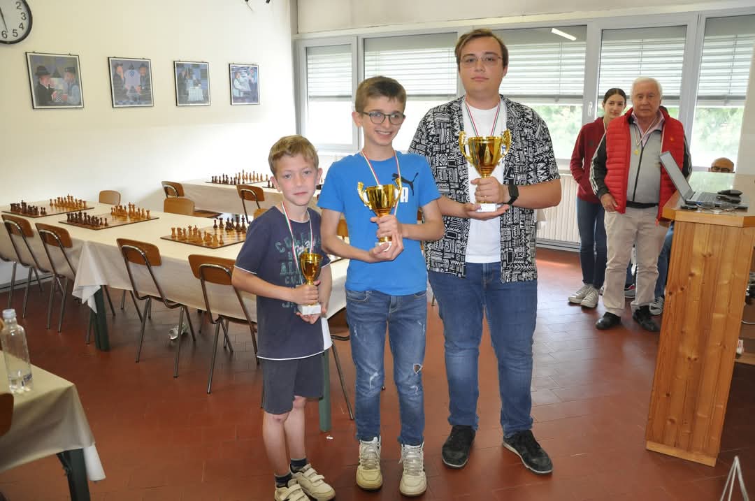 Tre bambini premiati con trofei in una sala, con tavoli e sedie sullo sfondo.