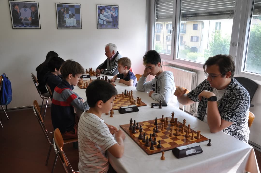 Un gruppo di bambini e adulti partecipa a un torneo di scacchi, seduti attorno a tavoli con scacchiere in un ambiente luminoso e accogliente.