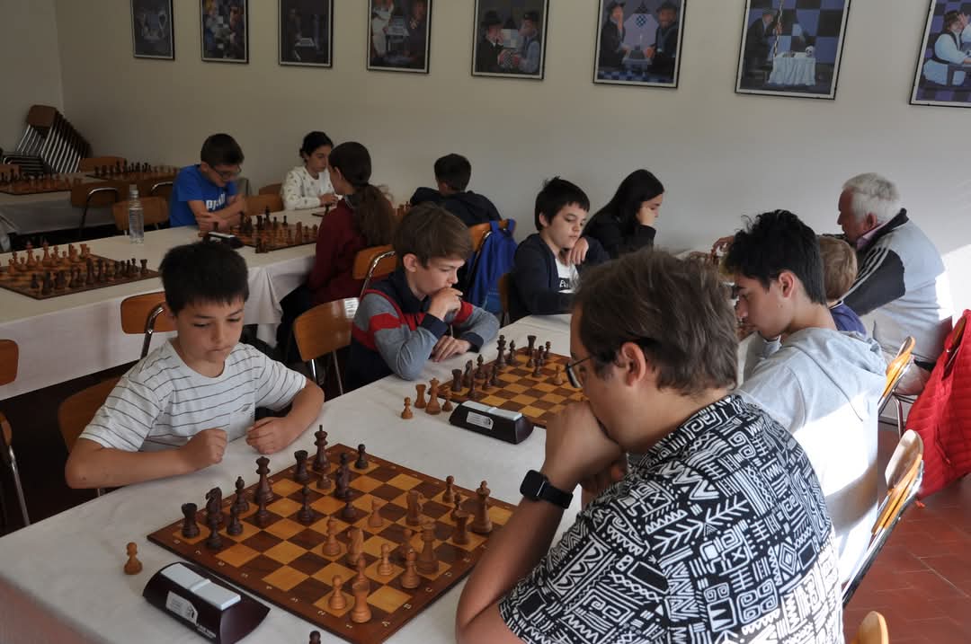 Bambini e adolescenti che giocano a scacchi in una sala con foto alle pareti.