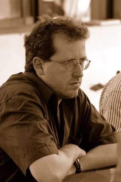 Un uomo con occhiali e capelli ricci, seduto in una posizione pensierosa, con le braccia incrociate, in un ambiente di gioco di scacchi.