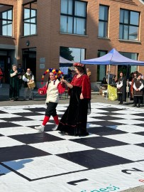 Two performers dressed in vibrant costumes walk on a large checkerboard during a festival, with people in the background observing.