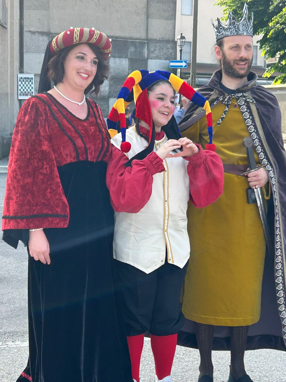 Three people dressed in medieval costumes, one in a red and black gown, another in a jester outfit with colorful hat and face paint, and the third in a king's attire with a crown.