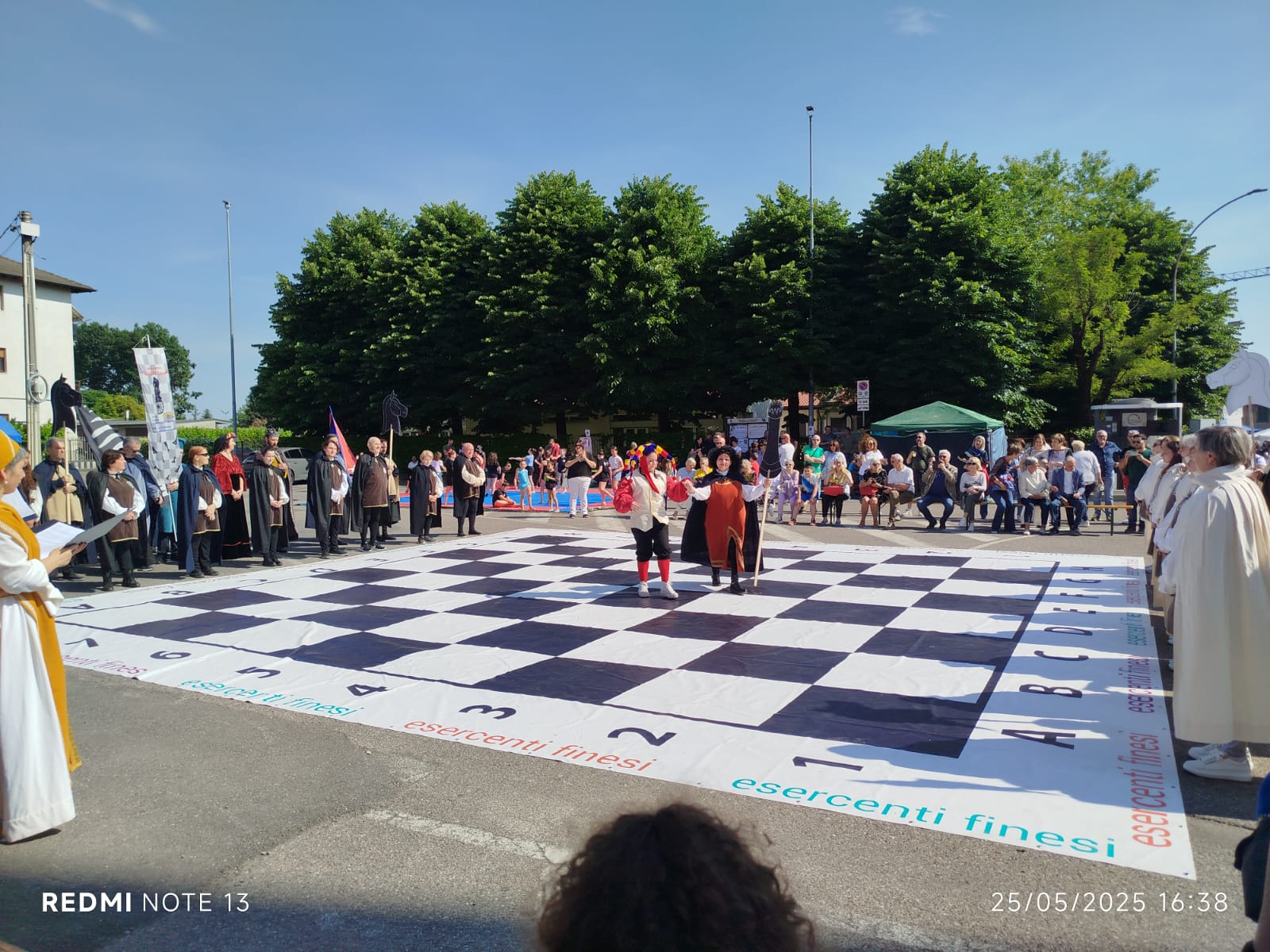 Outdoor chess tournament with a large checkered board and players in the center, surrounded by an audience.
