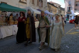 A group of three people dressed in historical costumes walking together on a street lined with market stalls.