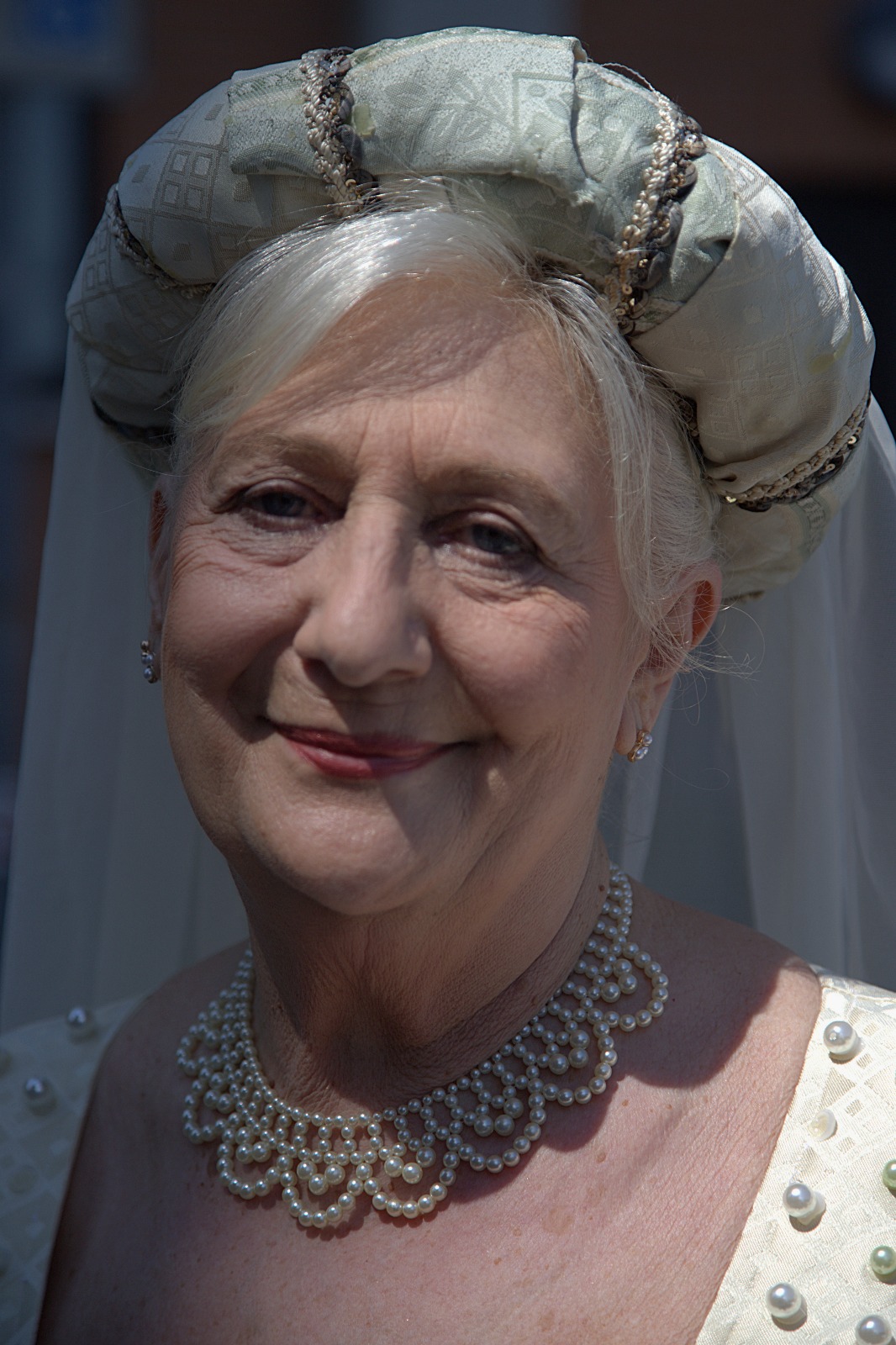 An elderly woman wearing a pearl necklace and a decorative headpiece, smiling warmly at the camera.