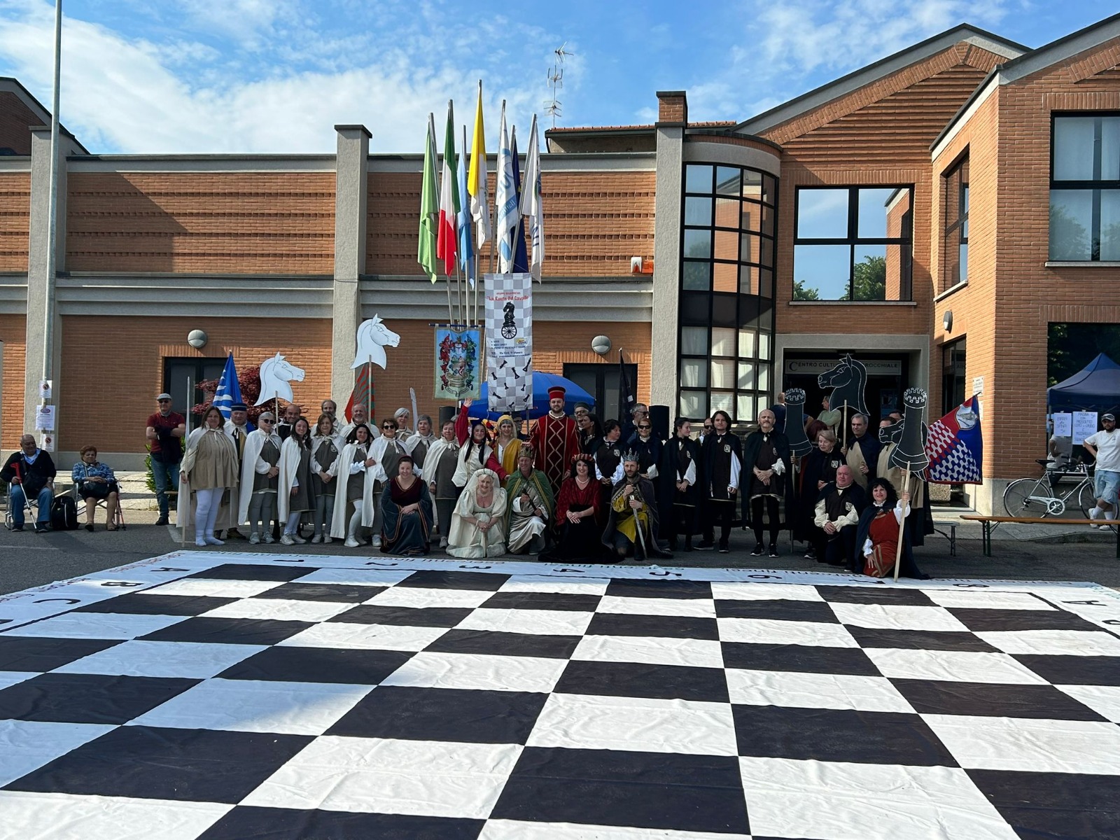 A group of people dressed in various costumes posing on a large chessboard outside a building, with flags and banners displayed in the background.