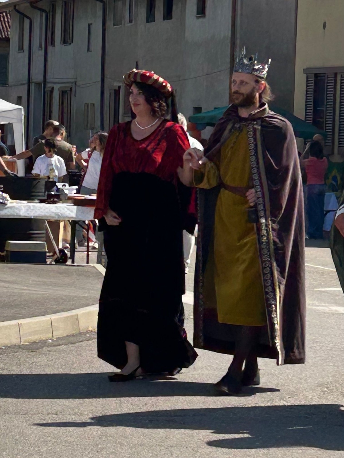 A man dressed as a king and a woman in a black dress with a red overlay walk together in a street, surrounded by people and market stalls.