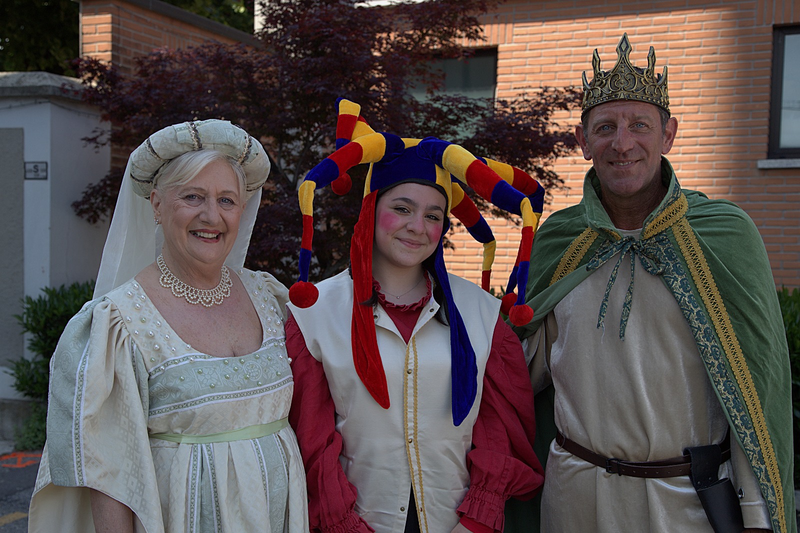 A woman in a white dress and elaborate necklace, a young woman in a jester costume with colorful hat, and a man in a crown and cloak pose together outdoors.