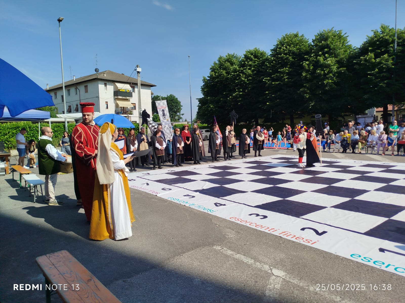 A large outdoor chessboard is set up with players about to start a game, surrounded by spectators and trees.