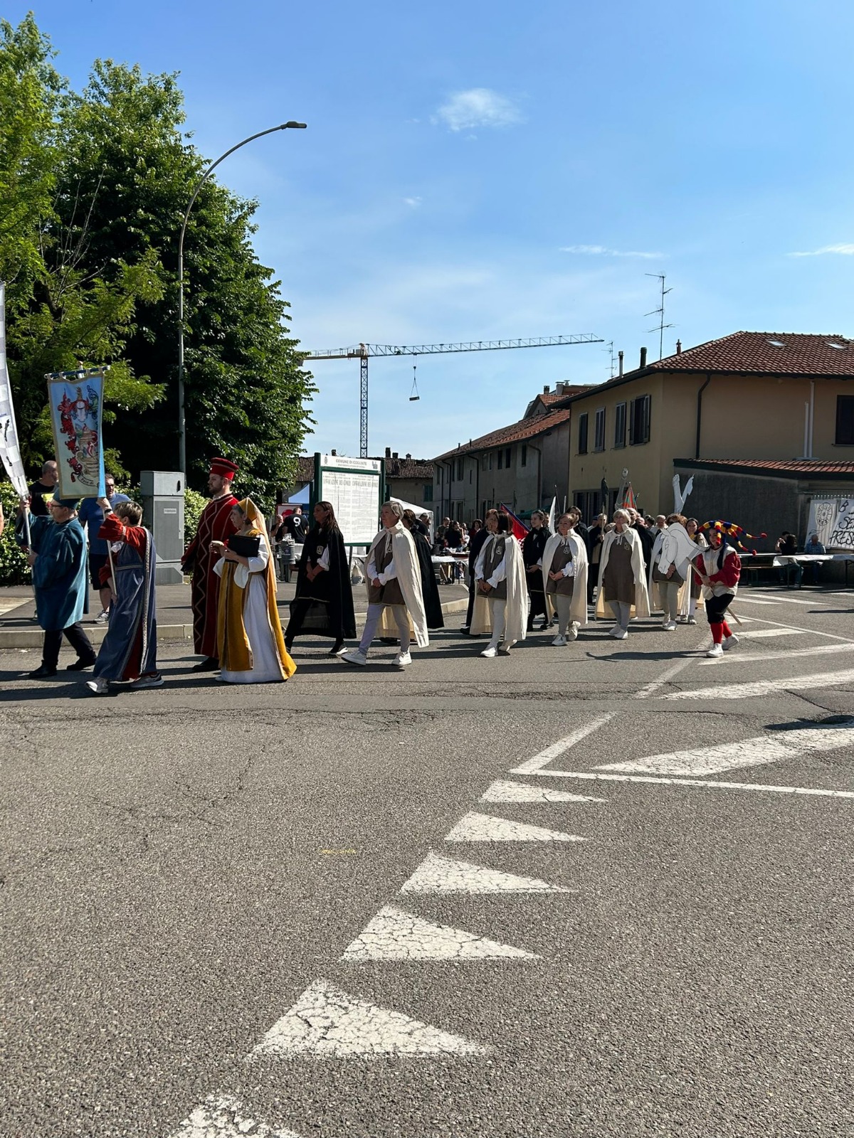 A group of people in costumes participating in a parade on a sunny day, walking along a street lined with buildings and trees.