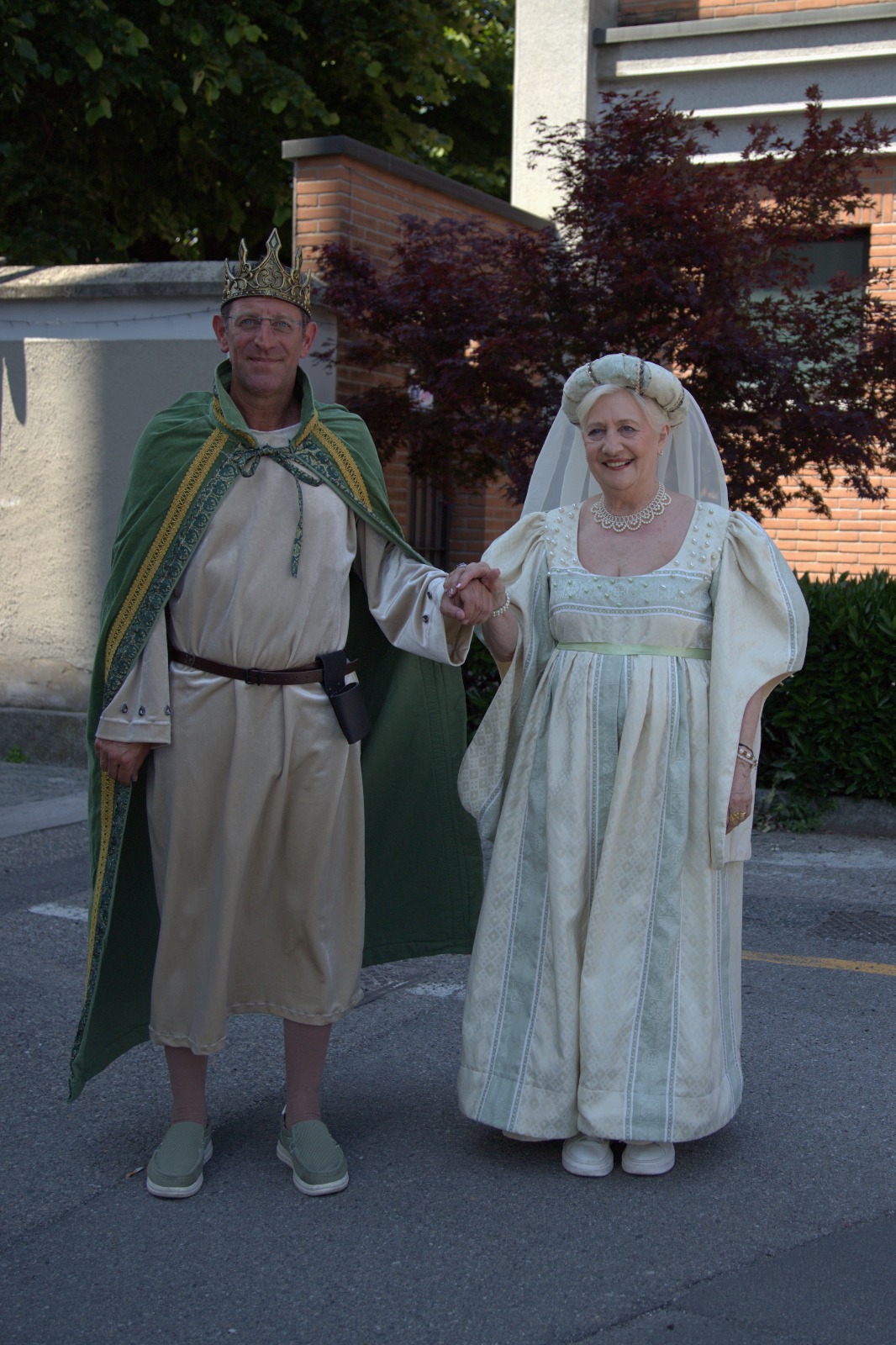 A man and a woman dressed in medieval-style costumes, standing hand in hand. The man is wearing a crown and a green cape, while the woman is in a light-colored gown with a veil.