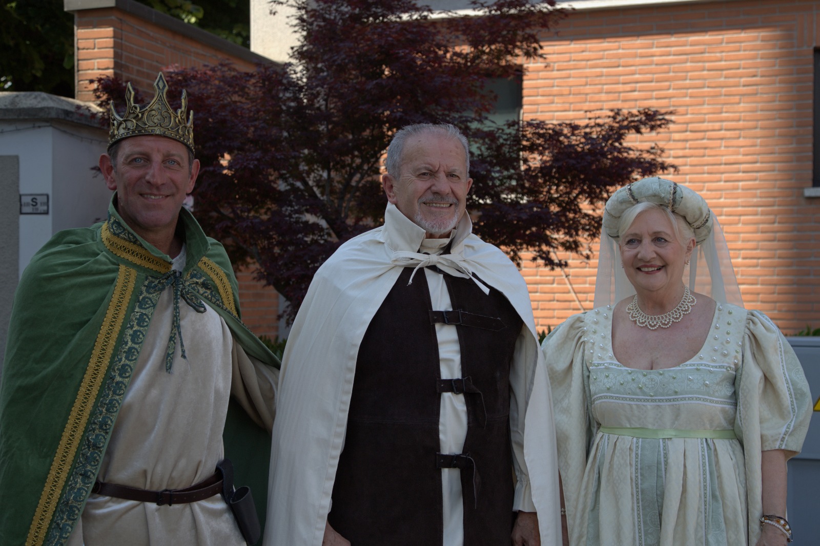 Three individuals in costumes; a man wearing a crown and green cape, a man in a white cloak with a dark robe underneath, and a woman in a light dress with a white veil, standing outdoors with trees in the background.
