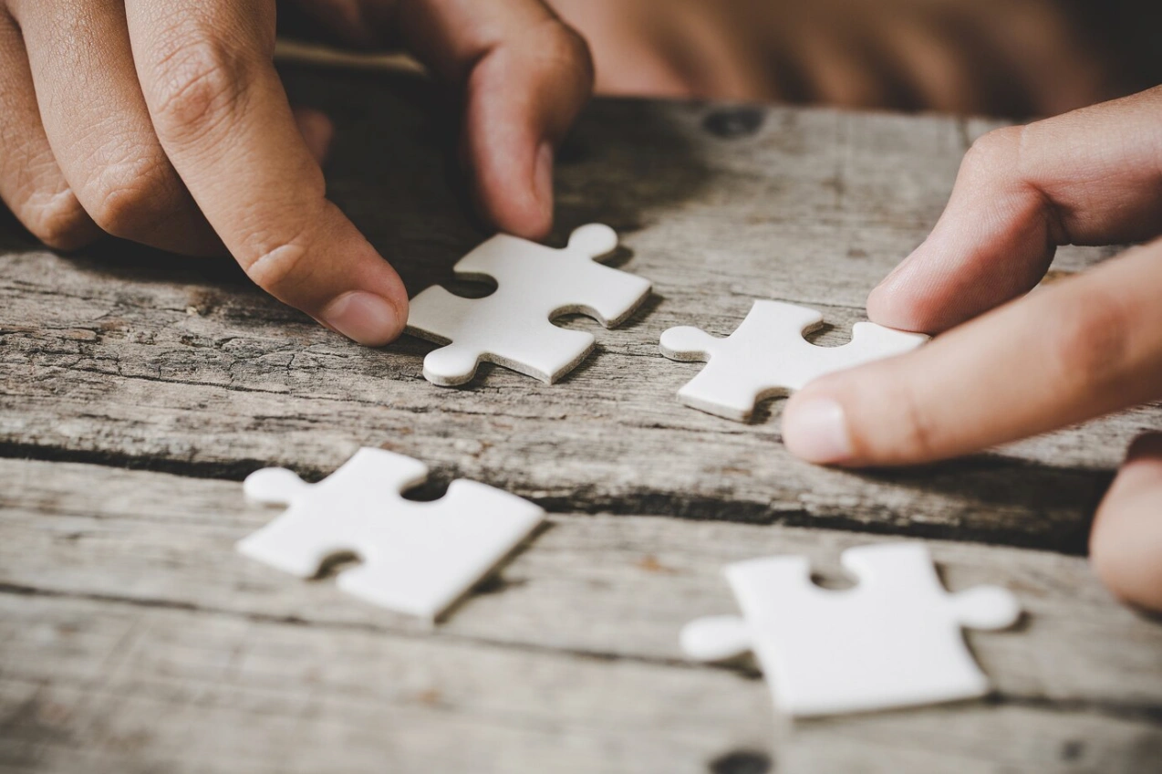 Due mani stanno maneggiando pezzi di puzzle bianchi su un tavolo di legno.