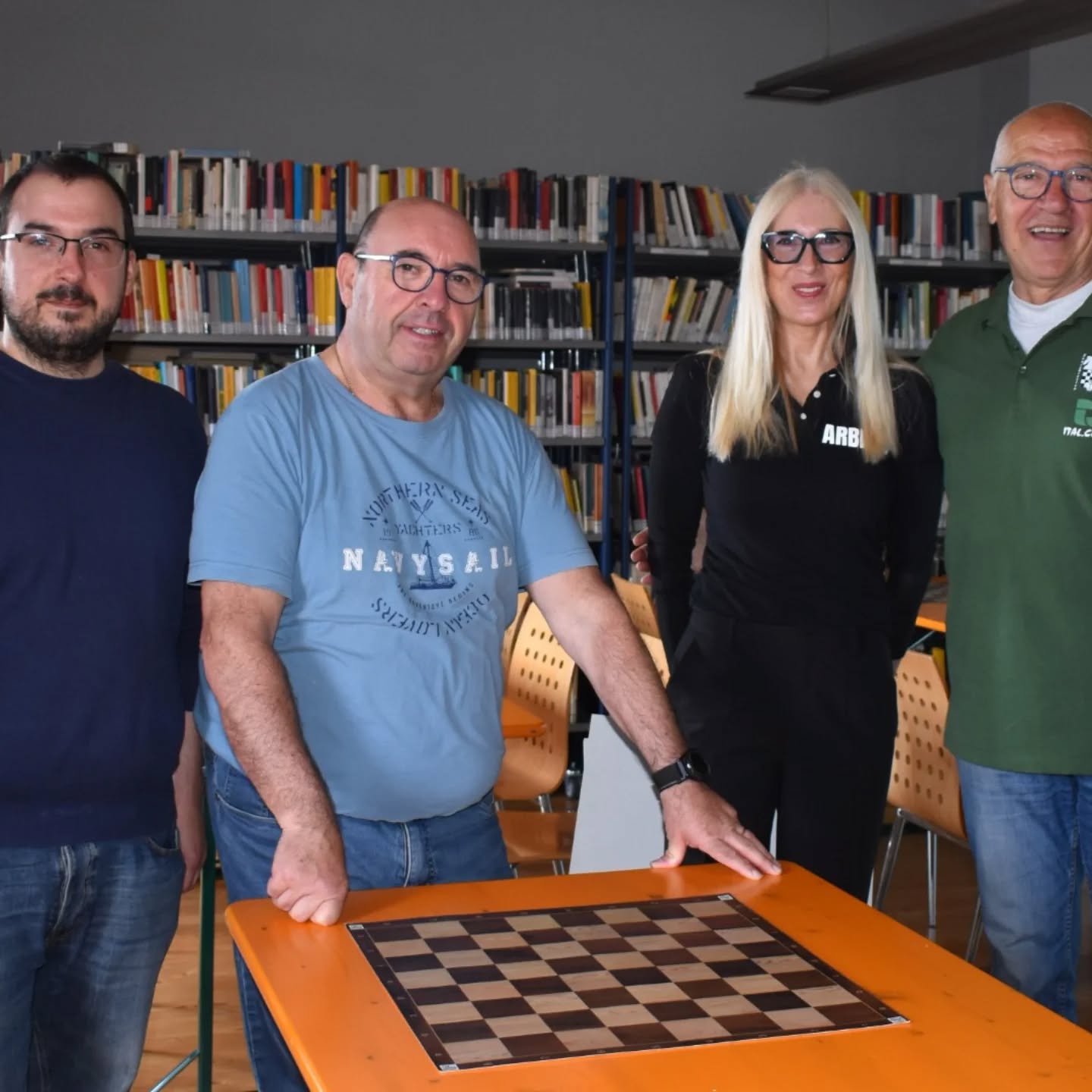 Un gruppo di quattro persone sorridenti si trova accanto a un tavolo con una scacchiera in una biblioteca. Alle spalle si vedono scaffali pieni di libri.