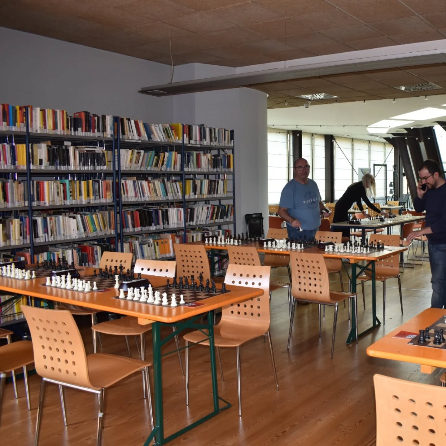 Interno di una biblioteca con scacchiere disposte su tavoli e scaffali pieni di libri, mentre alcuni partecipanti si preparano per un torneo di scacchi.