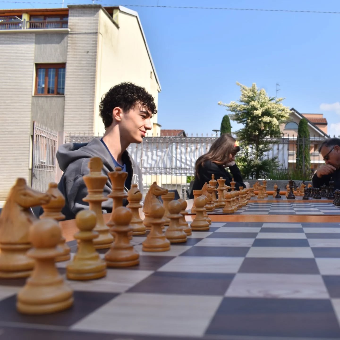 Due persone stanno giocando a scacchi all'aperto, con una scacchiera in legno e pezzi scolpiti ben visibili in primo piano.