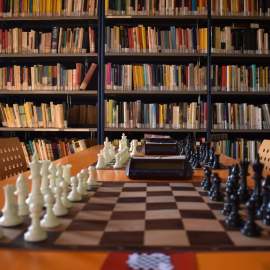 Una scacchiera di legno con pezzi bianchi e neri, situata di fronte a una libreria con libri colorati sullo sfondo.