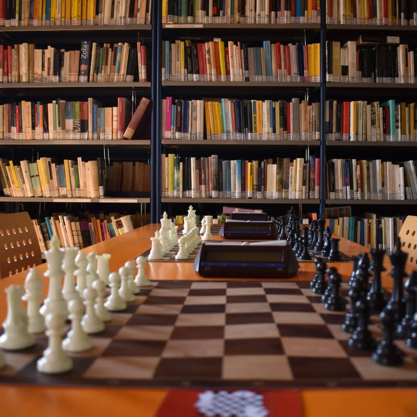 Scacchiere bianche e nere disposte su un tavolo in una biblioteca, con uno sfondo di scaffali pieni di libri.