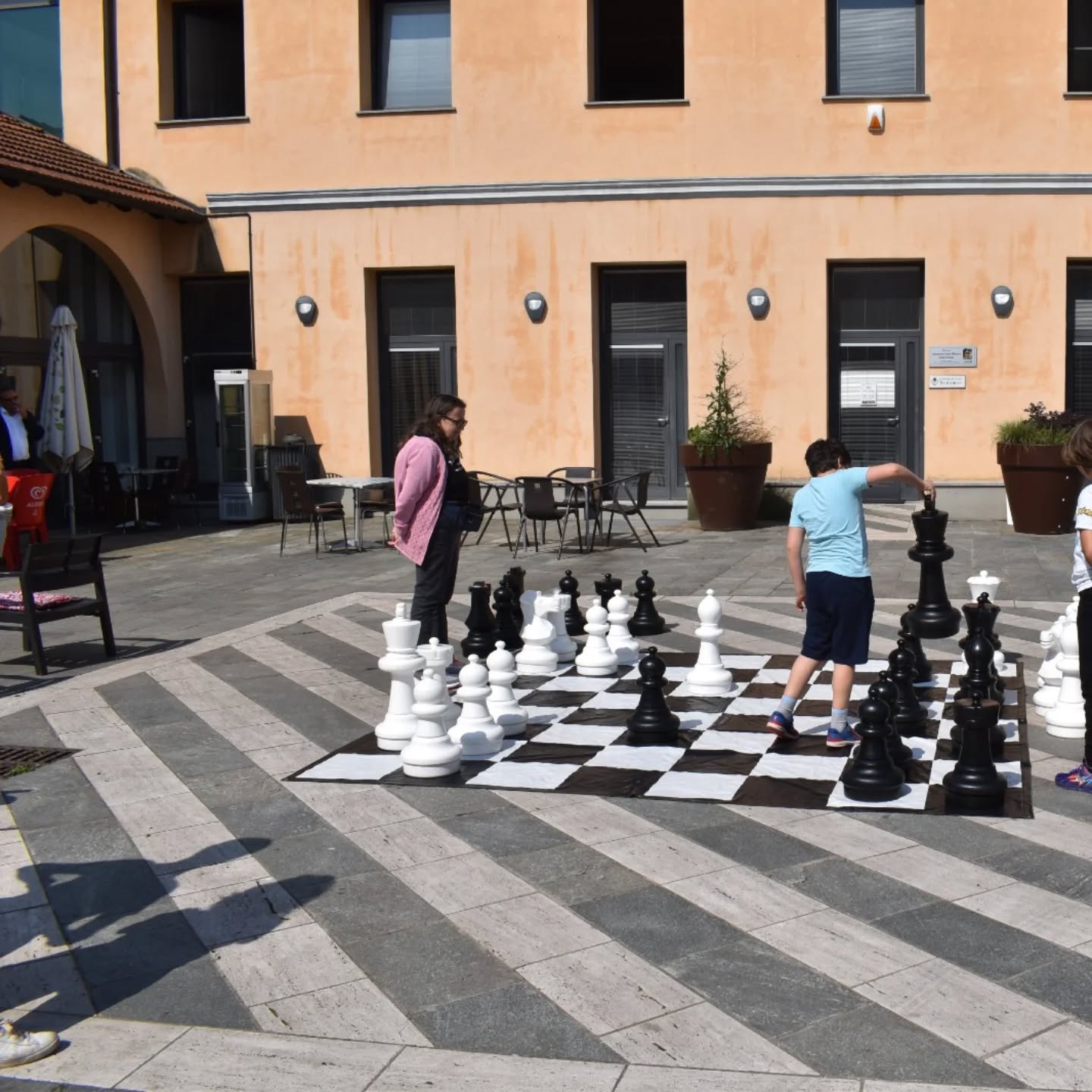 Giocatori che partecipano a una partita di scacchi su un grande scacchiere in un'area esterna, con edifici sullo sfondo.