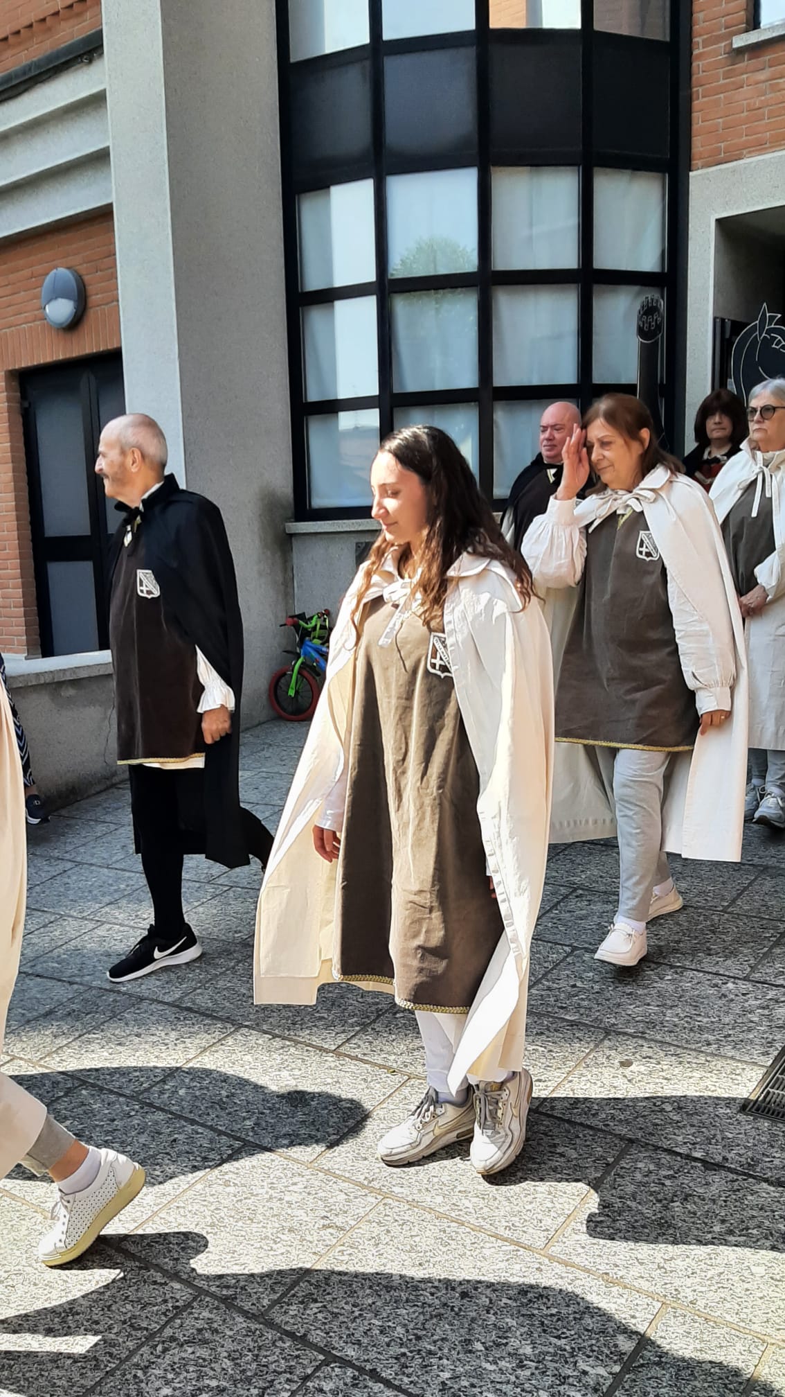 A group of individuals dressed in brown and white garments is walking outside a building, with some participants wearing capes and different types of footwear.
