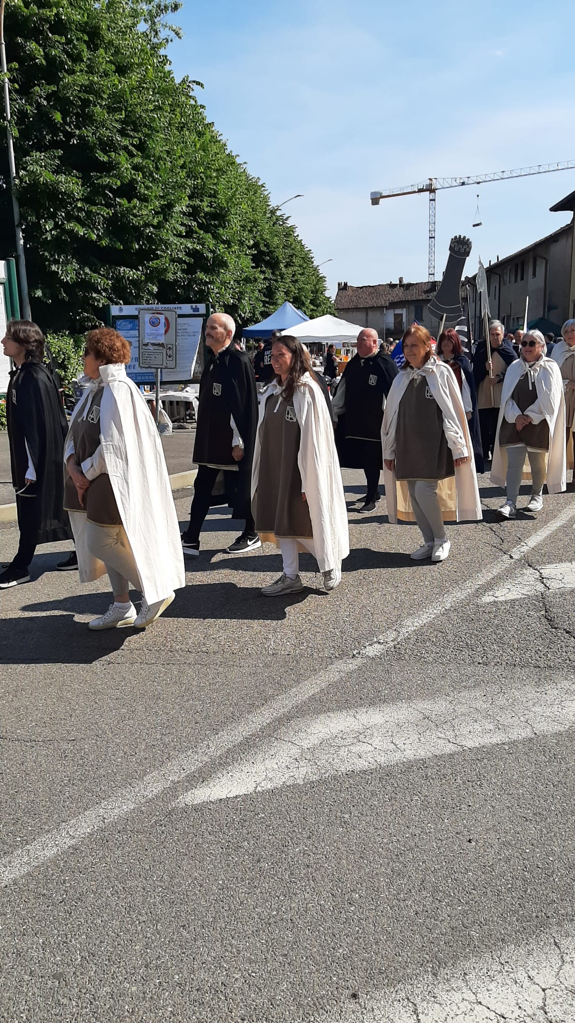 A group of people wearing capes and cloaks participating in a procession on a street, with trees and buildings in the background.
