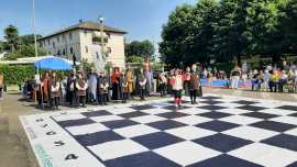 A large outdoor chessboard with people gathered around, observing a game in progress. The setting includes a building and trees in the background under clear skies.