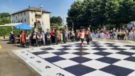 A large outdoor checkerboard with people gathered around, some watching a performance on the board, and a building in the background.