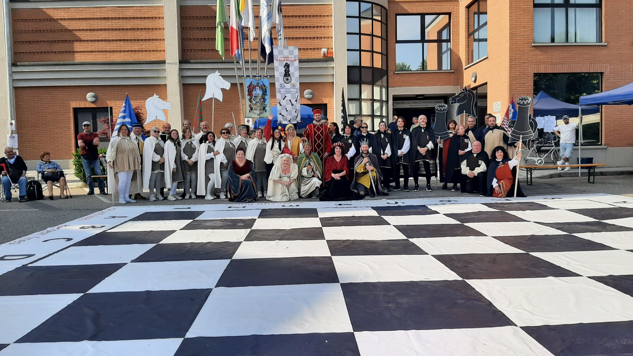Group of people in historical costumes gathered around a large chessboard in an outdoor setting.