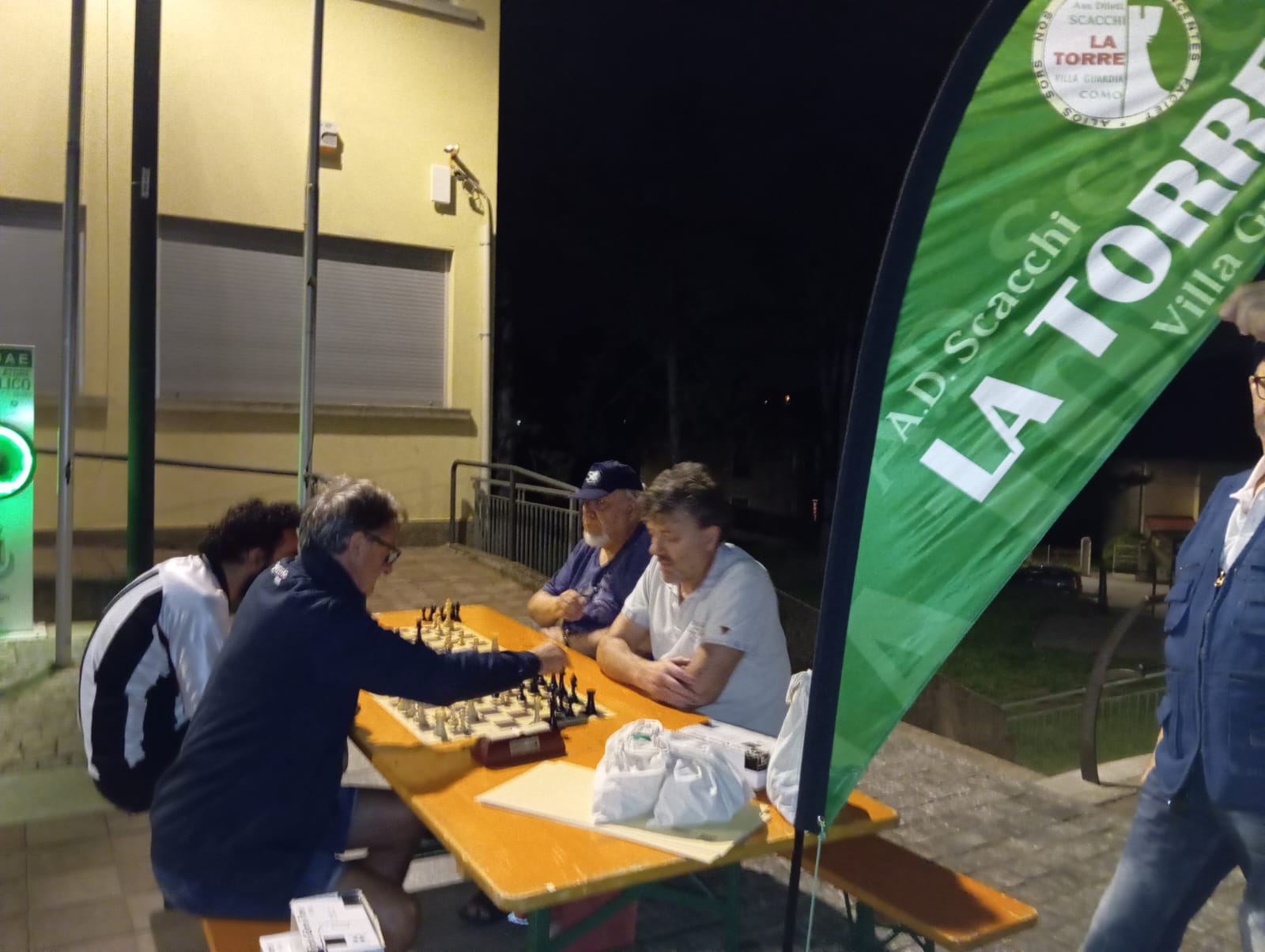Persone che giocano a scacchi attorno a un tavolo all'aperto durante un evento serale, con una bandiera verde che pubblicizza un'attività.