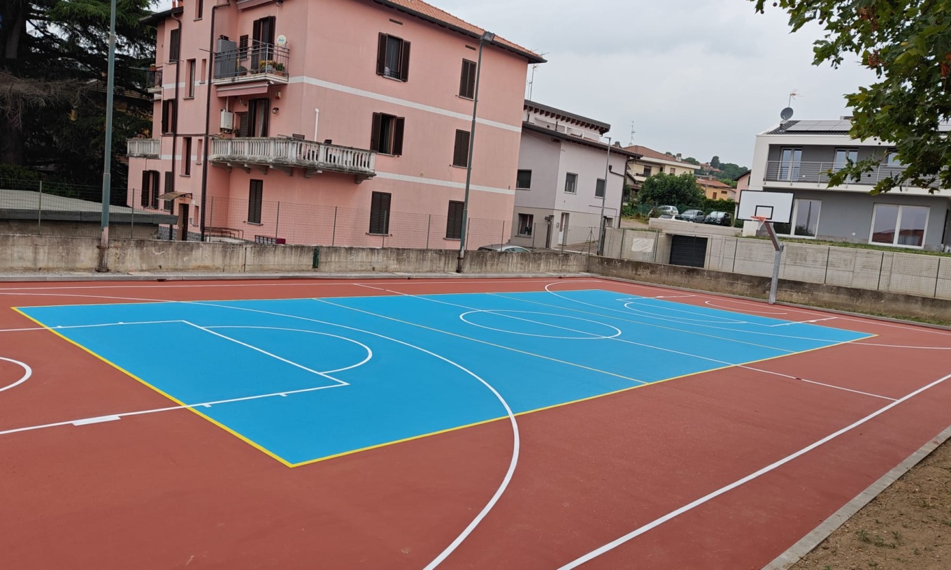 A basketball court with a bright blue surface, surrounded by buildings and trees.