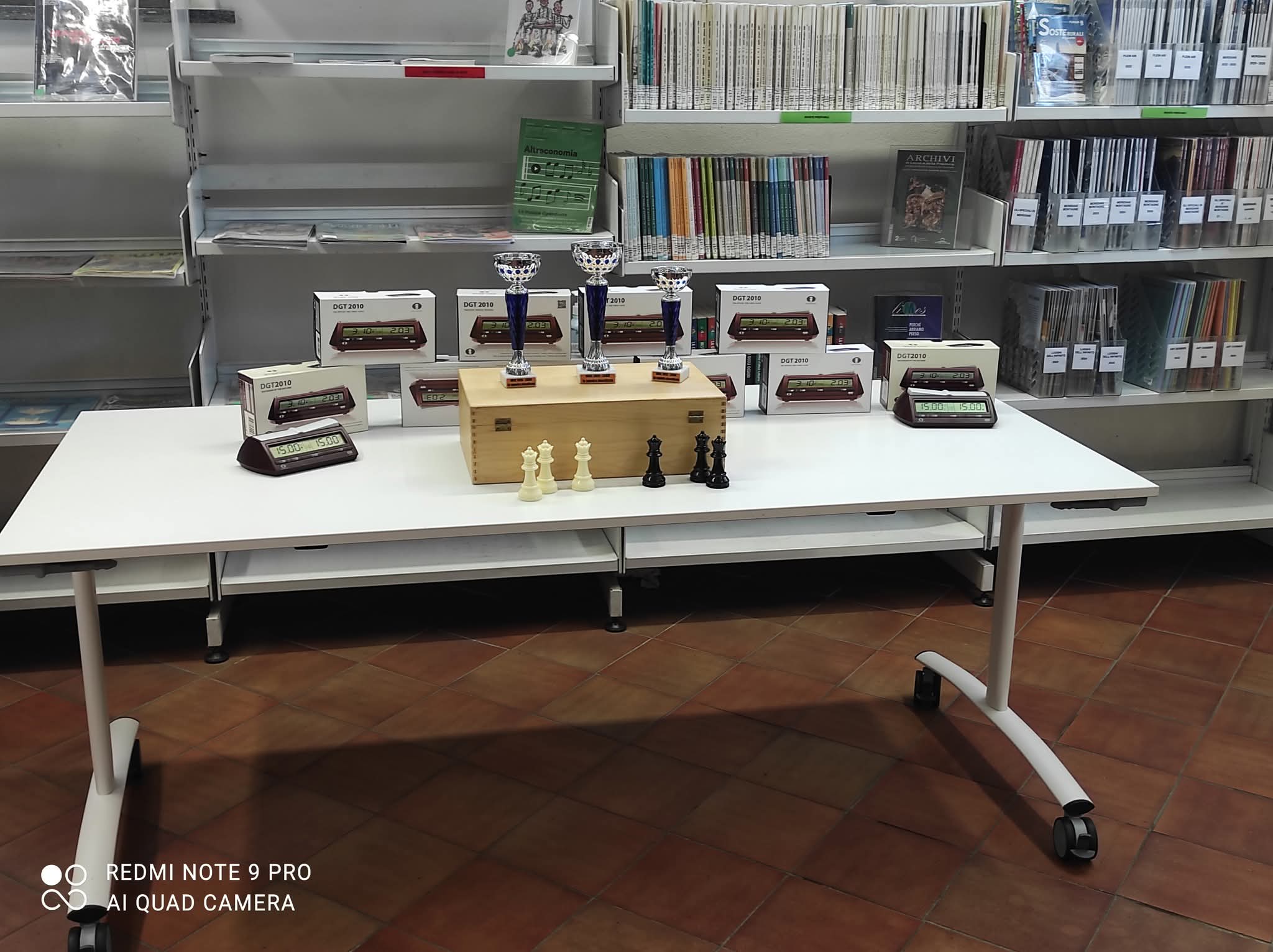 Una tavola bianca con trofei, medaglie e pezzi di scacchi, allestita all'interno di una biblioteca.