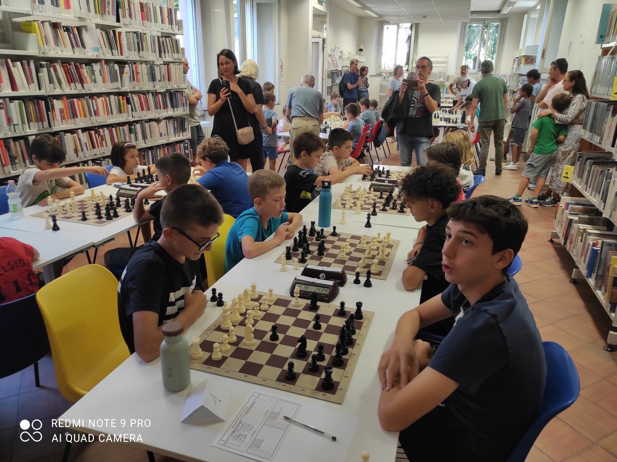 Bambini che partecipano a un torneo di scacchi in una biblioteca, concentrati sui loro giochi con pezzi di scacchi e tavoli di legno.