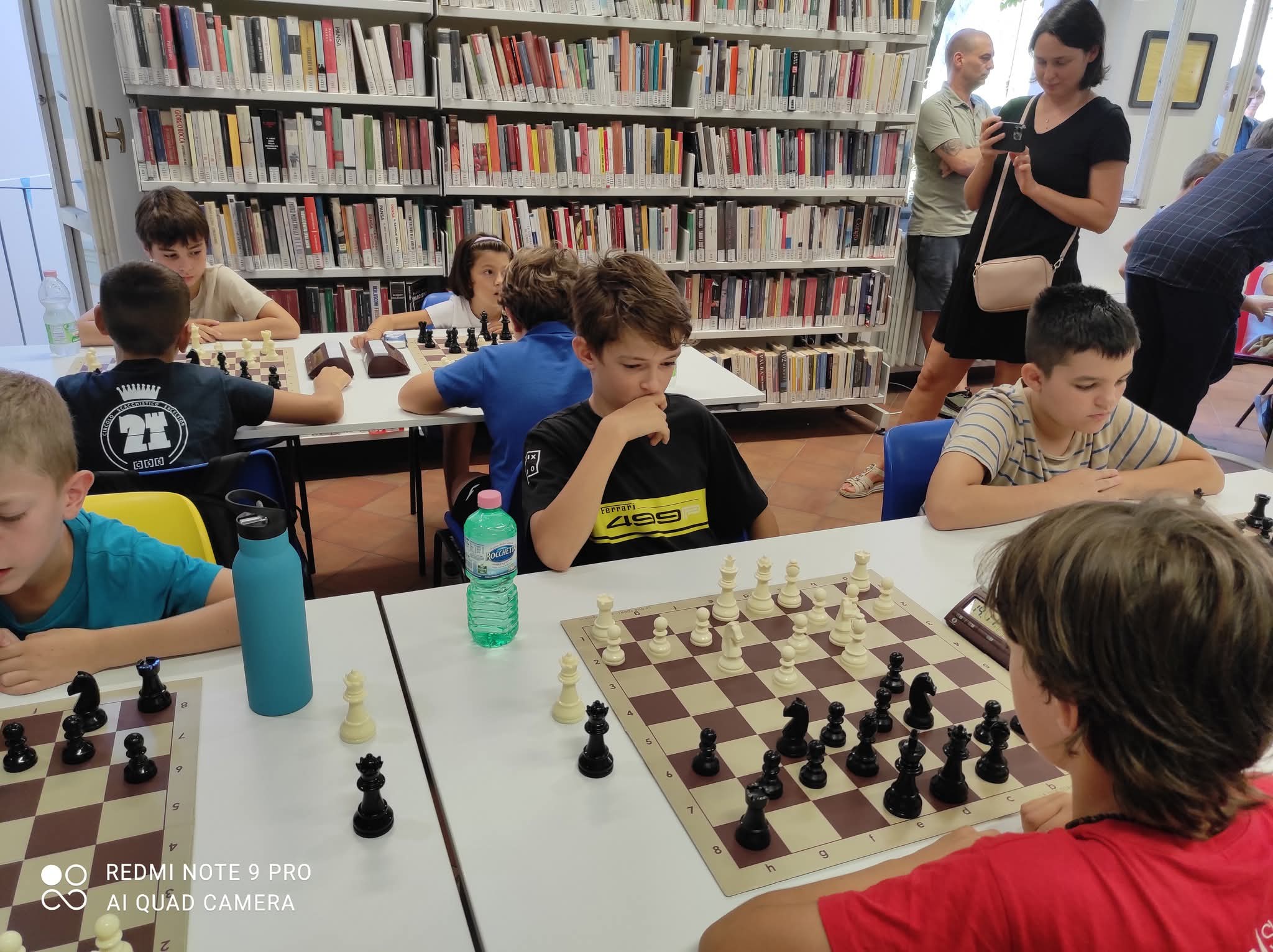 Un gruppo di ragazzi gioca a scacchi in una sala di una biblioteca, con scacchiere e pezzi bianchi e neri, mentre adulti osservano in background.