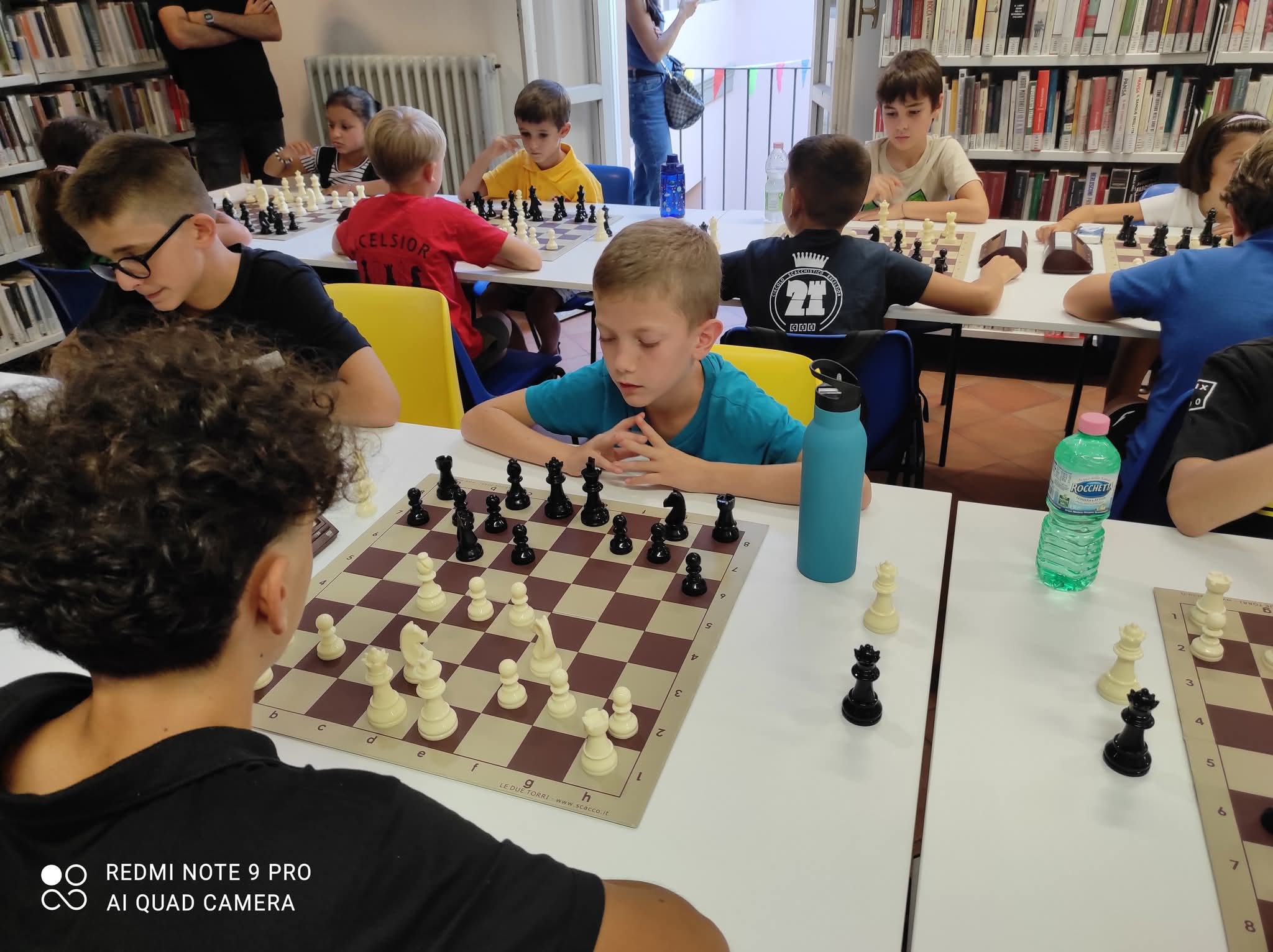 Giocatori di scacchi durante una partita in biblioteca, con tavoli allestiti e bambini concentrati sul gioco.