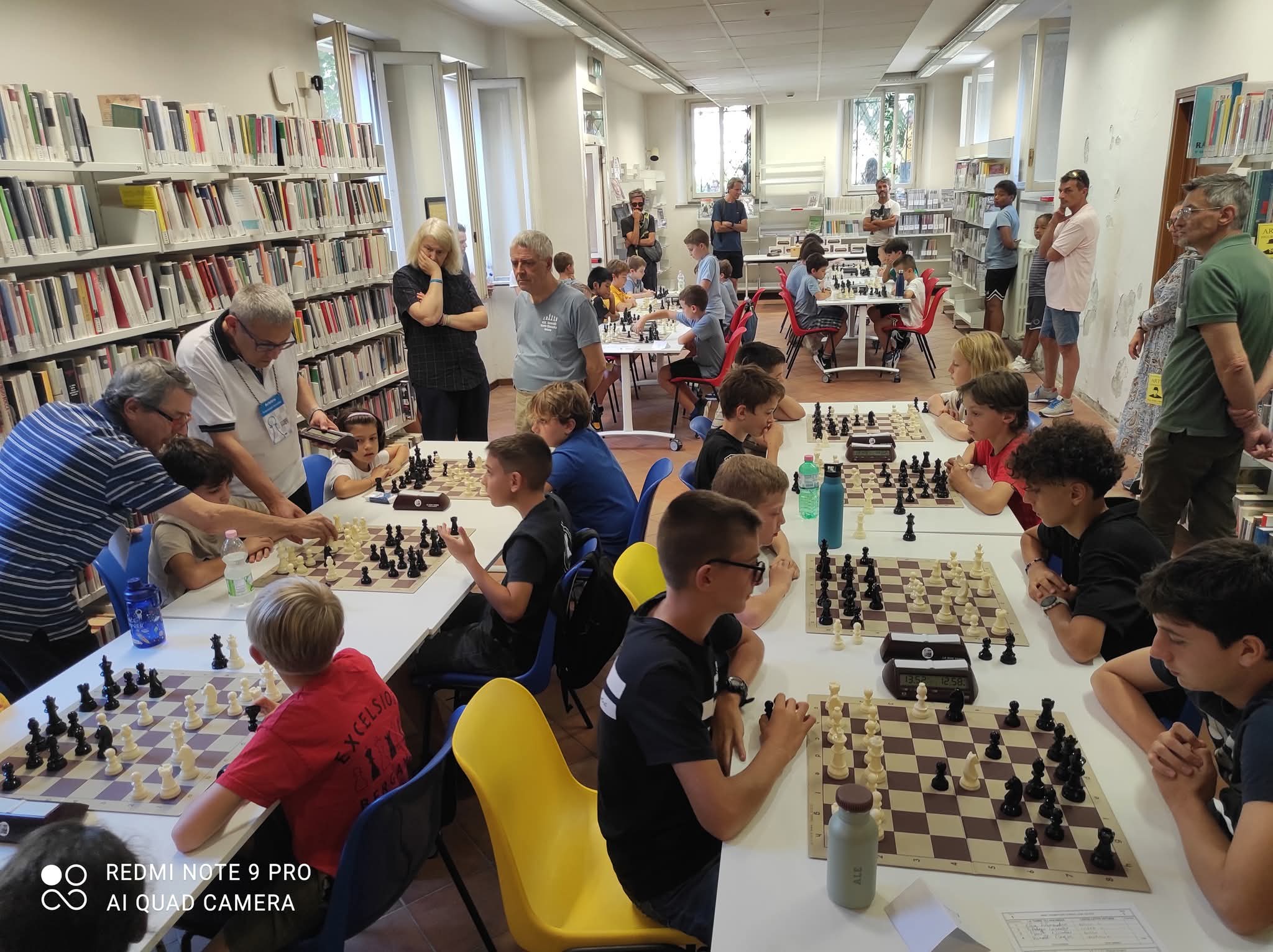 Giovani giocatori di scacchi in competizione a un torneo nella biblioteca civica di Lecco, con diversi tavoli da gioco e partecipanti concentrati sulle loro partite.