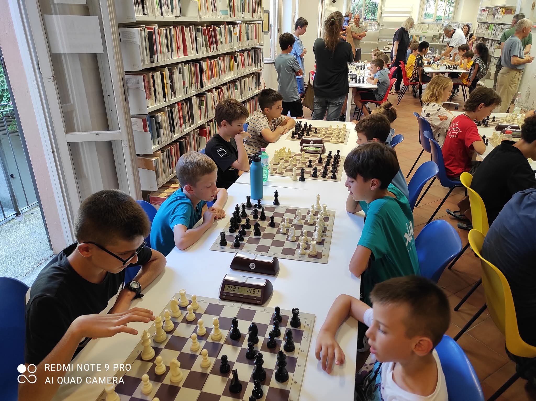 Un gruppo di bambini gioca a scacchi in una biblioteca, con diverse scacchiere e un ambiente vivace attorno a loro.