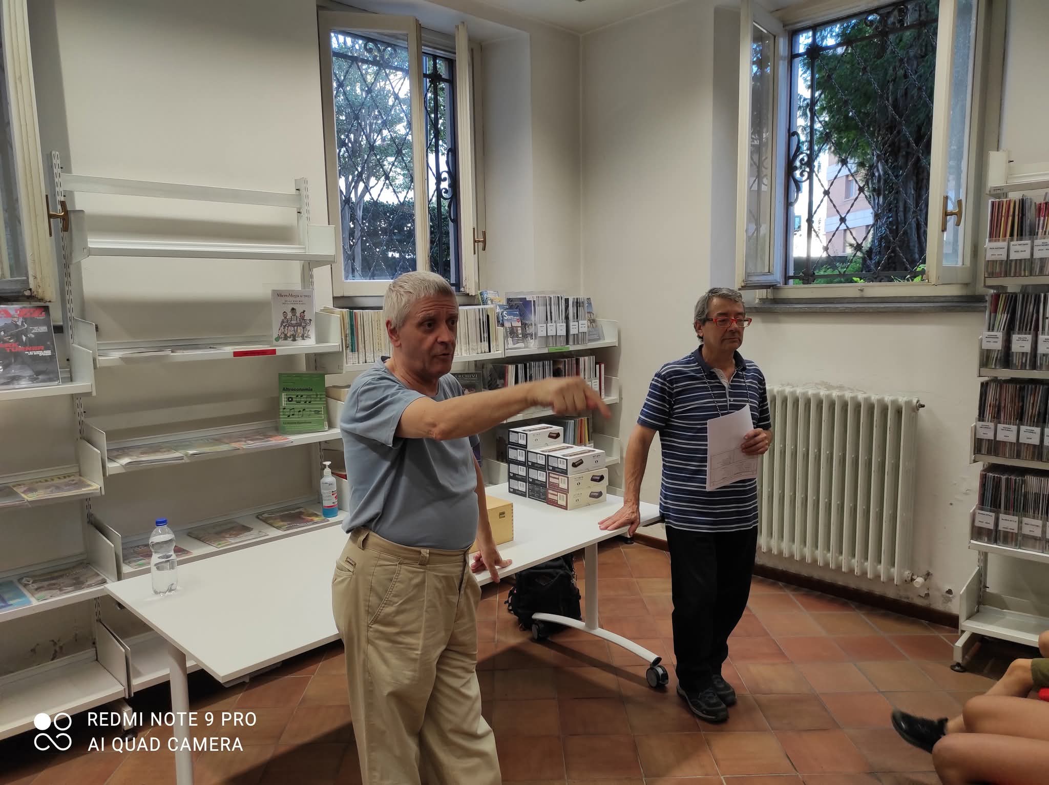 Due persone che parlano in una biblioteca, una con un foglio in mano e l'altra che gesticola.