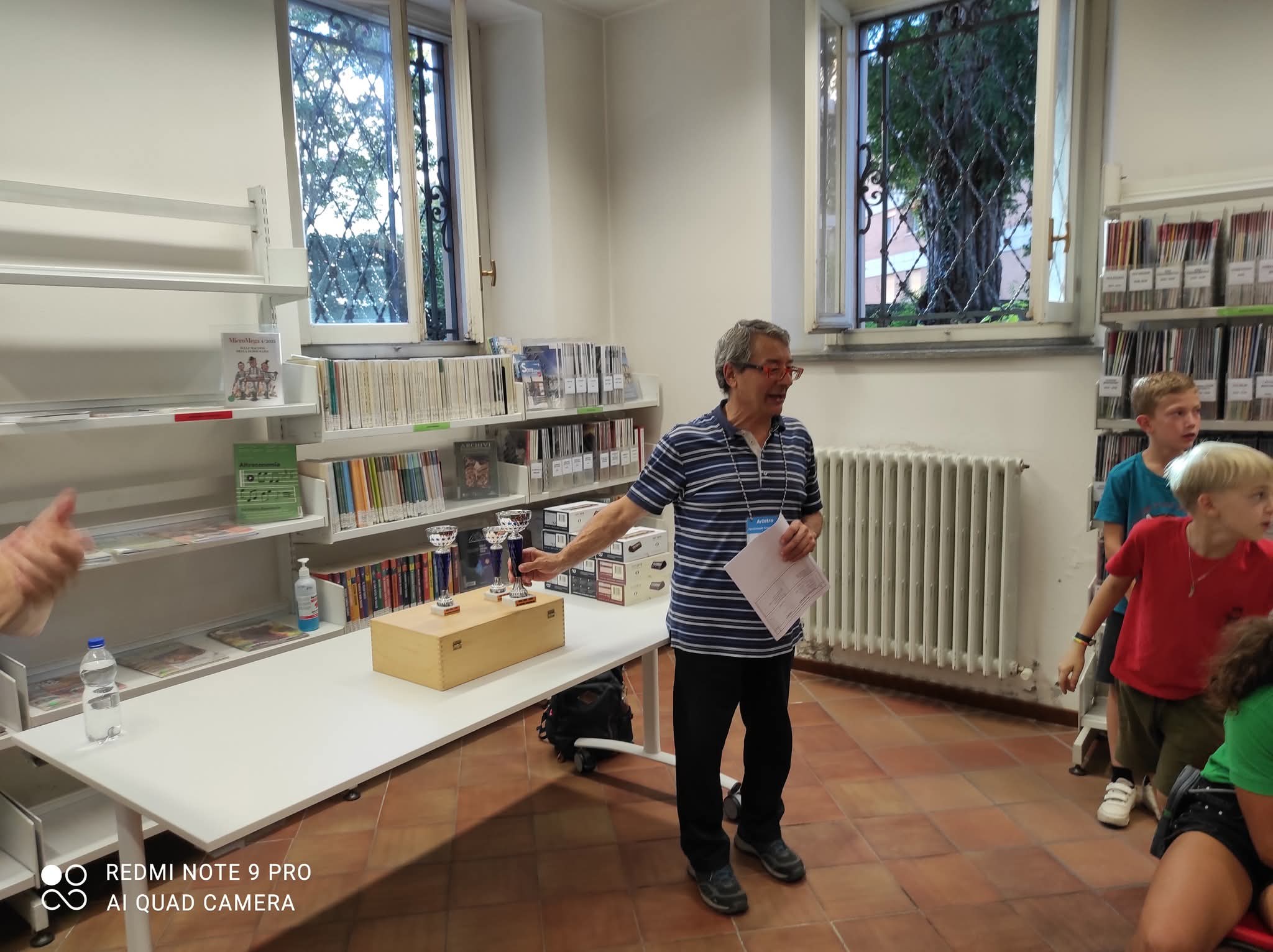 Un istruttore spiega le regole del torneo di scacchi nella biblioteca civica di Lecco, circondato da giovani partecipanti entusiasti.