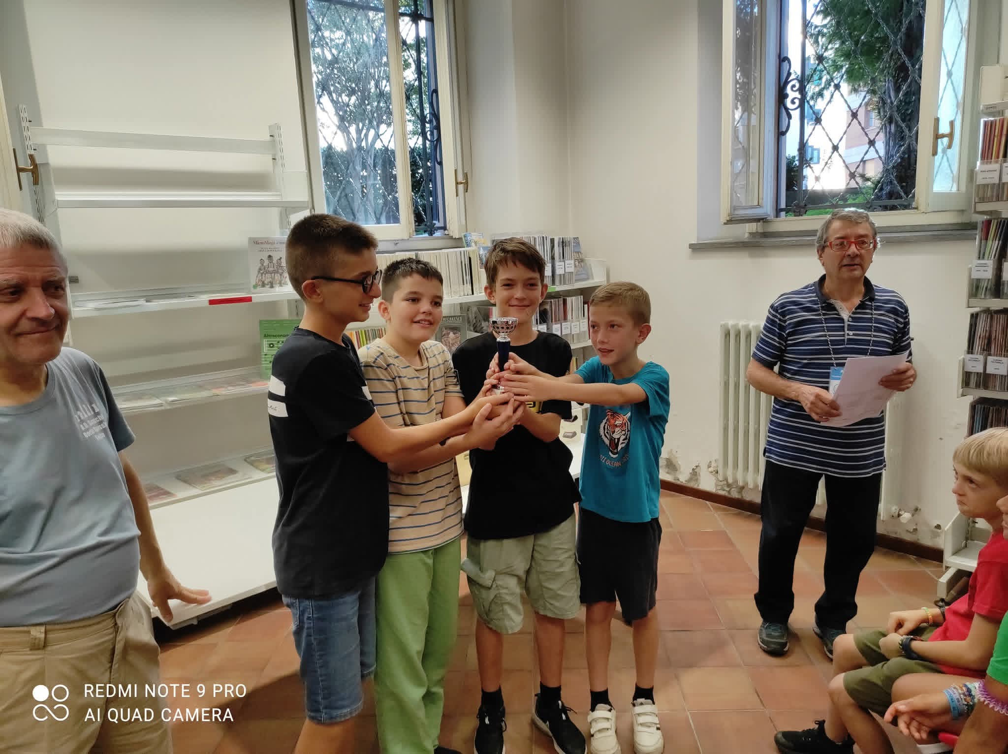 Un gruppo di ragazzi celebra la loro vittoria mentre ricevono un premio, con un adulto che legge un documento sullo sfondo.