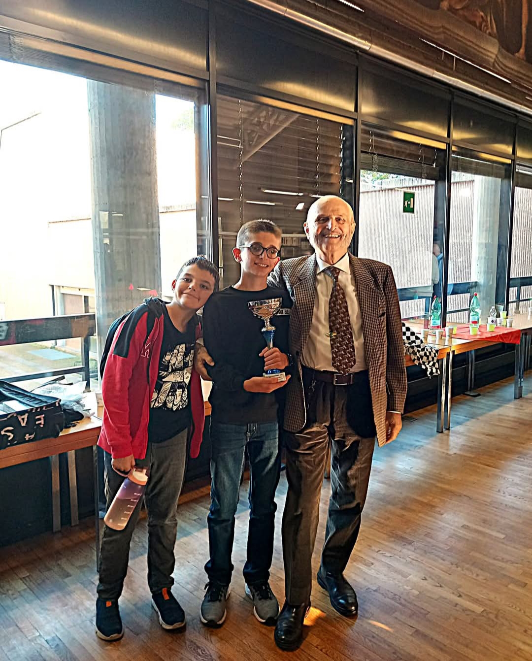 Un giovane atleta con trofeo al centro, affiancato da due persone in un ambiente di festa, con finestre panoramiche sullo sfondo.