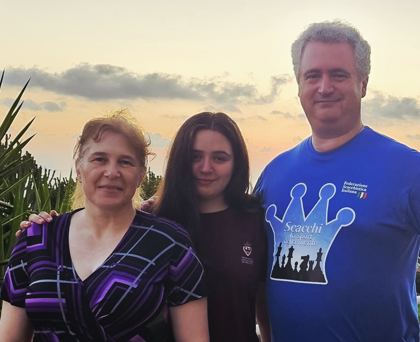 Un uomo e una donna sorridenti posano con una giovane donna, con il mare e il tramonto sullo sfondo, mentre si trovano all'aperto.