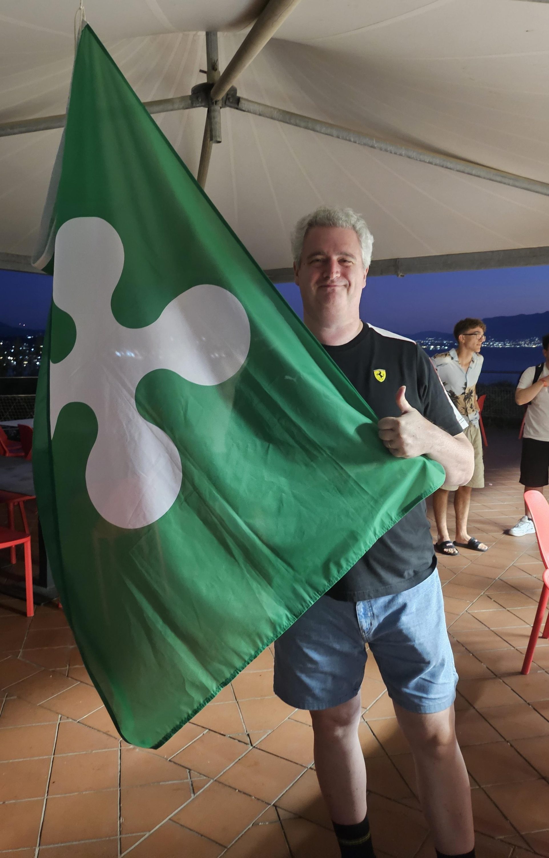 Un uomo sorride mentre tiene in alto una bandiera verde con un simbolo bianco, all'interno di un'ambientazione di festa all'aperto.