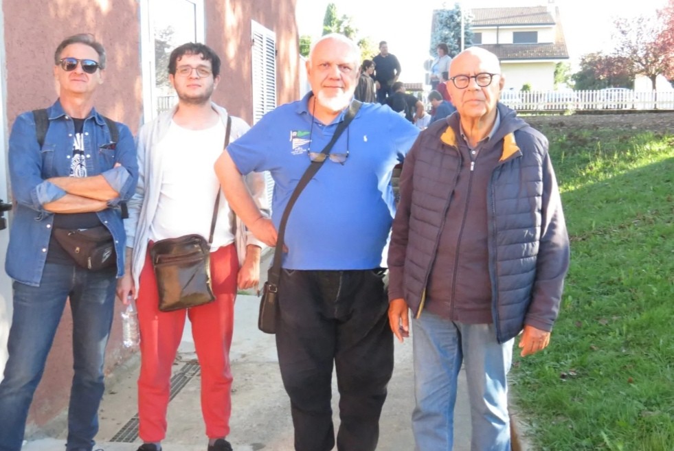 Quattro uomini sorridenti posano per una foto all'aperto, con un edificio e un prato sullo sfondo.