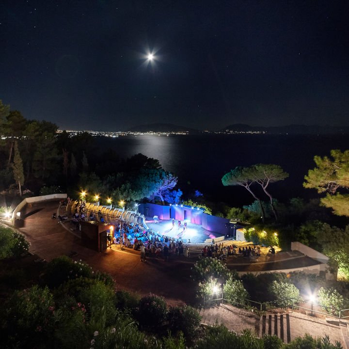 Vista panoramica di un evento serale all'aperto, con un pubblico riunito attorno a un palco illuminato. La scena è ambientata in un giardino con alberi e una vista sul mare sotto un cielo notturno stellato.