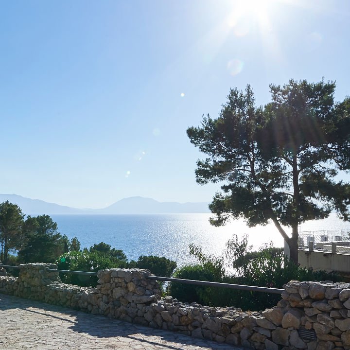 Vista panoramica di un mare calmo con una costa montuosa sullo sfondo, illuminata da una luce solare brillante. In primo piano, un paesaggio con alberi e una ringhiera di pietra.