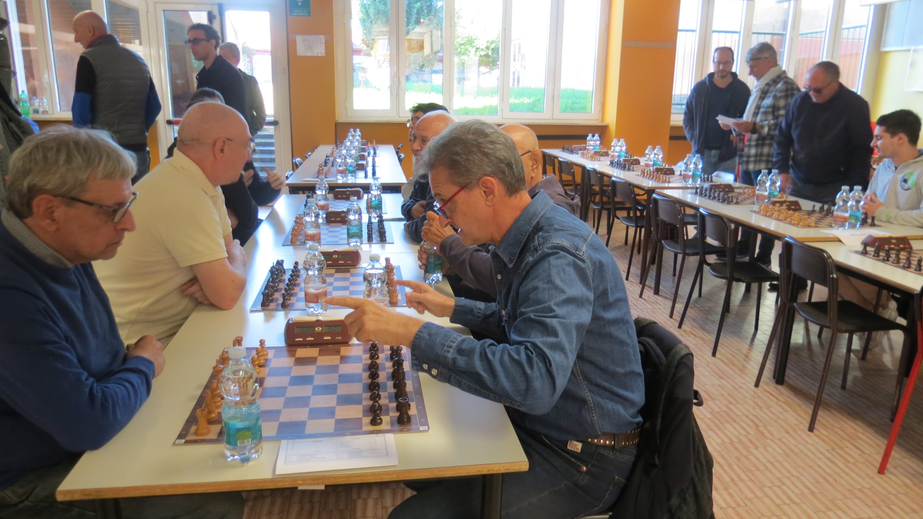 Two men playing chess at a table in a community hall, with other players in the background.