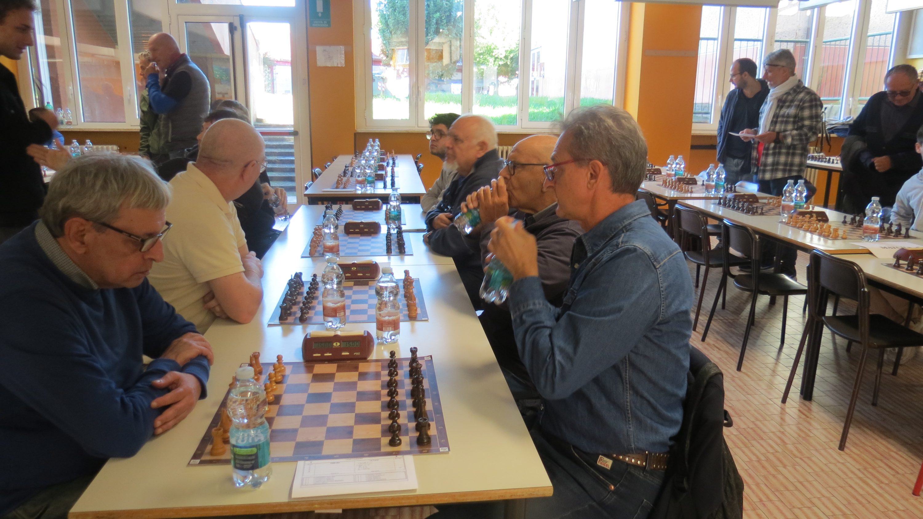 A group of people engaged in a chess game, seated at tables in a well-lit room with large windows.