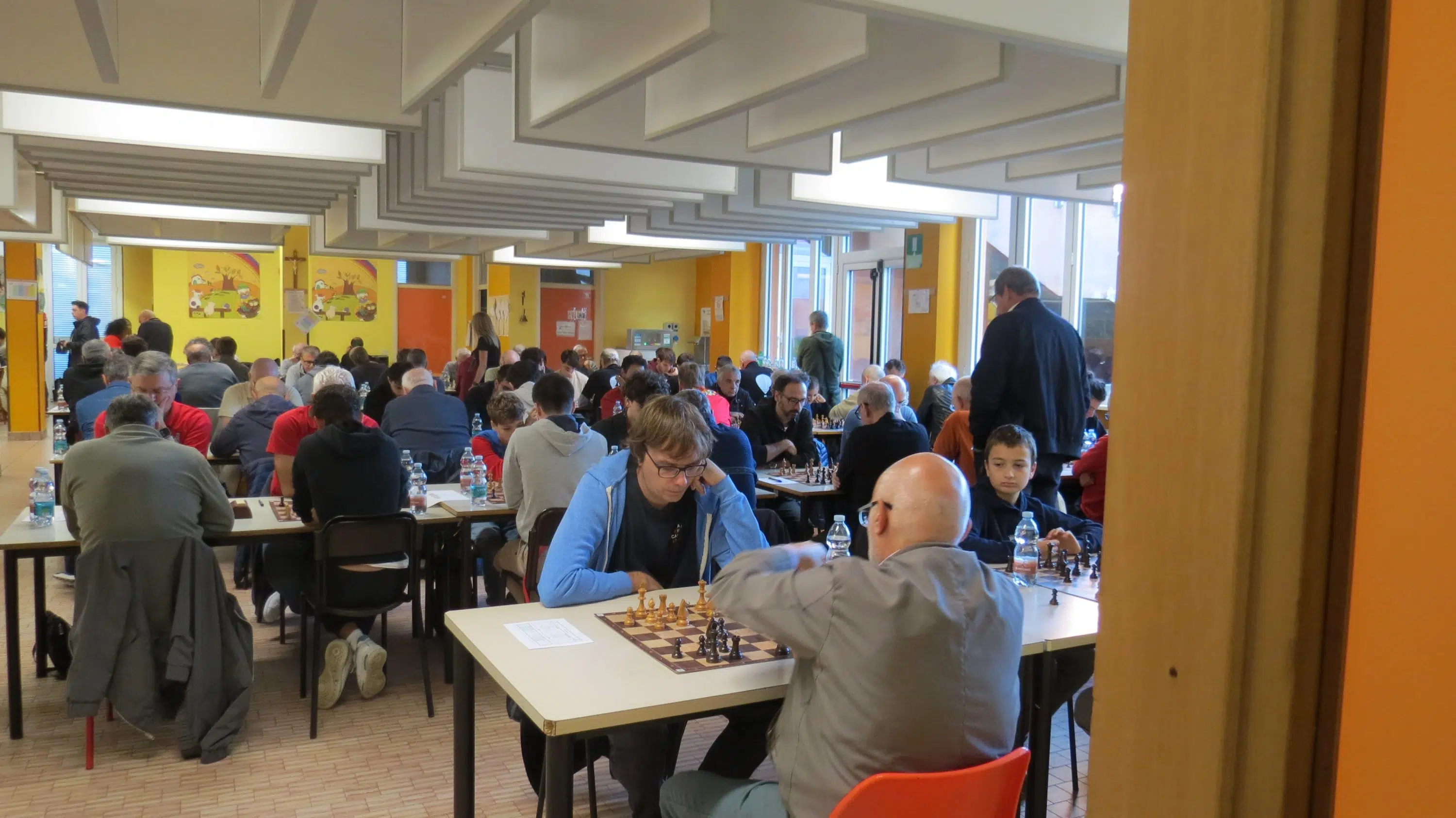 A crowded chess tournament taking place indoors, with participants engaged in games at tables surrounded by spectators.
